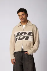 Campera de punto con cierre frontal completo, cuello polo y diseño boxy. Presenta una franja horizontal en el pecho con un estampado gráfico en color oscuro sobre fondo beige.