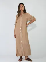 Vestido largo de lino color tostado, tipo camisero con cuello clásico, cierre frontal completo con botones y costuras en contraste. Presenta mangas largas con presilla para arremangar y aberturas laterales en el bajo.