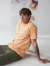 Camiseta de manga corta color naranja claro, con cuello redondo y un bolsillo en el pecho. Presenta un efecto de teñido en prenda que le da un aspecto ligeramente desgastado.