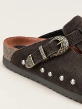 Zueco de gamuza marrón oscuro, forrado en cuero vacuno, con tira ajustable con hebilla metálica decorativa y tachas plateadas alrededor de la suela.