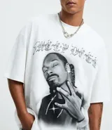 Remera blanca de algodón con cuello redondo, mangas cortas y corte oversized. Presenta un estampado frontal en blanco y negro del rapero Snoop Dogg, con letras góticas en la parte superior.