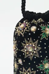 Cartera tipo bolso de mano color negro, confeccionada en terciopelo con bordados de estrellas y aplicaciones de pedrería brillante. Cuenta con correas trenzadas de cordón negro y cierre superior.