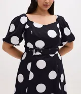 Vestido midi de algodón con estampado de lunares blancos sobre fondo negro. Presenta escote en V, mangas cortas abullonadas, cintura ajustada con cinturón de hebilla y falda con tajo lateral.