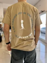 Remera color camel con cuello redondo y mangas cortas, con diseño gráfico estampado en la espalda que incluye un pájaro con sombrero de copa y la frase circular "Focus on finding joy in the journey".