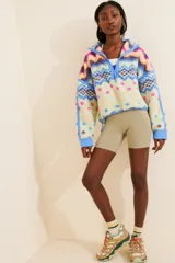 Buzo de polar con diseño estampado geométrico en tonos verde, blanco y naranja. Presenta cuello alto con cierre frontal parcial, mangas largas con puños y cordón ajustable en el ruedo.