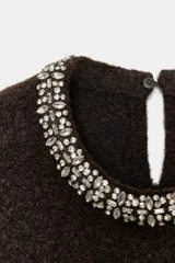 Jersey de punto color marrón oscuro, de corte recto y manga larga. Presenta un cuello redondo decorado con abalorios joya y una abertura en la espalda con cierre de botón.