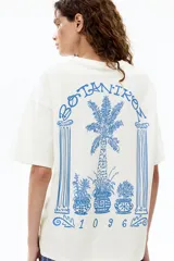 Remera oversized de algodón blanco, con cuello redondo acanalado, mangas cortas con hombros caídos y pequeño estampado azul en el pecho de un jarrón con flores y el número 1096.
