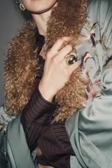 Anillo maxi de metal con engaste ovalado. El engaste tiene un borde exterior trenzado en tono oscuro y un borde interior liso en tono dorado, sosteniendo una piedra grande facetada de color transparente.