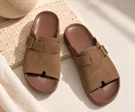 Sandalia plana tipo chinela de gamuza marrón, con tira ancha en el empeine, hebilla metálica lateral y diseño anatómico.