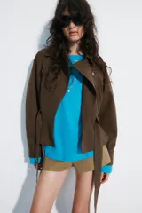 Cazadora trench corta color marrón, confeccionada en mezcla de algodón, con cuello solapa, cierre cruzado con botones, bolsillos de vivo en el delantero y cinturón ajustable del mismo tejido.