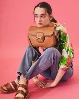 Cartera tipo bandolera de cuero con textura grabada, color beige. Presenta un detalle de herraje metálico dorado en la solapa frontal y placa con logo de la marca. Incluye correa ajustable para llevar al hombro o cruzada.