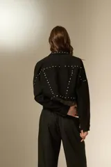 Chaqueta de jean negra con corte holgado, cuello camisero, cierre frontal con botones y detalle de strass y tachas metálicas distribuidas en el frente y mangas.