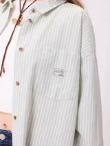 Camisa de corte oversize con diseño a rayas verticales en tonos azul y blanco. Presenta cuello clásico, cierre frontal con botones y un bolsillo en el pecho con detalle de etiqueta.