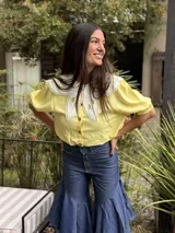 Camisa de lino amarillo claro, con mangas cortas abullonadas y un amplio cuello Peter Pan blanco con detalles bordados y calados. Cierre frontal con botones.