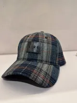 Gorra con diseño a cuadros tartán en tonos gris, azul marino y marrón, con un parche de cuero sintético negro en el frente que lleva grabado el logo de la marca 'AIRE'.