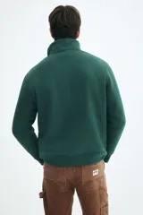 Buzo de tejido pesado color verde oscuro, con cuello alto, medio cierre metálico y hombros caídos. Presenta puños y bajo en punto acanalado.