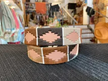 Cinturón de cuero en tono tostado con bordado a mano de hilos encerados en tonos marrones y beige con motivos geométricos, y hebilla metálica rectangular.