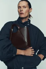 Bolso de mano color marrón oscuro con doble asa y asa extraíble para usar como bandolera. Presenta detalles de pespuntes en el cuerpo y cierre con botón imantado.