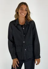 Chaqueta de jean negra con cuello camisero, cierre frontal con botones metálicos, bolsillos de parche en el pecho y ruedo desflecado.