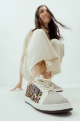 Zapatillas urbanas de mujer, color blanco, con puntera de gamuza gris claro, detalles en cuero blanco, panel lateral con estampado de leopardo y logo de corona en cuero marrón oscuro. La suela es de goma gruesa blanca y negra.