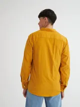 Camisa de manga larga color mostaza confeccionada en pana fina, con bolsillo en el pecho, botones de pasta, bordado en el ruedo y etiqueta de marca en la tapeta de los botones.