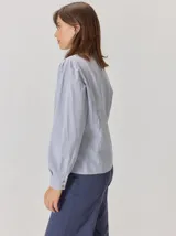 Camisa de denim con silueta levemente entallada, cuello bebé y mangas largas con sutil frunce en los hombros. Presenta costuras curvas decorativas con pespuntes en contraste y cierre frontal con botones.