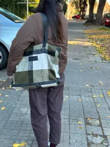 Bolso tipo tote de gran capacidad con diseño de cuadros en tonos verde oliva, blanco y negro. Presenta detalles en cuero negro en la base y en las correas de hombro, además de un parche de cuero con logo en el frente. Incluye cierre superior y bolsillo interior.