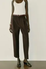 Pantalón tobillero de tiro medio, color beige, con pinzas delanteras y bolsillos frontales. Incluye un cinturón fino de cuero marrón con hebilla metálica dorada y cierre frontal con cremallera, botón interior y gancho.