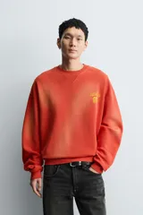 Sudadera de corte relajado, color naranja con efecto tie-dye/deslavado, cuello redondo y manga larga. Presenta un estampado frontal con texto y un trébol de cuatro hojas en amarillo.