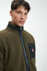 Buzo de abrigo verde militar, confeccionado en tejido sherpa, con cuello alto, cierre frontal con cremallera, bolsillo en el pecho con cierre y logo bordado.