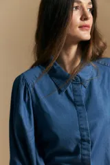 Camisa de denim azul con cuello camisero, cierre frontal de botones y mangas largas abullonadas con hombros fruncidos.