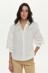 Camisa blanca de algodón con mangas abullonadas, cuello camisero y bordados geométricos y florales en tonos celestes y amarillos en el frente y las mangas.