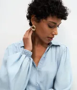 Camisa de denim liviano confeccionada en lyocell, con cuello camisero, cierre frontal de botones y mangas largas abullonadas con puños elásticos ajustados.
