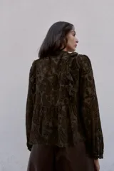 Blusa de manga larga con cuello redondo, confeccionada en tejido semitransparente con estampado paisley en tonos marrones oscuros. Presenta detalles de volantes y apliques de terciopelo en el frente y hombros, con cierre frontal de botones forrados.