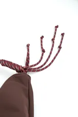 Bolso tipo morral color marrón de diseño curvo, con manija de cuerda trenzada en tonos burdeos y detalles de nudos en los extremos.