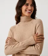 Suéter de tejido de punto color beige con cuello alto, mangas con corte raglán y puños y ruedo acanalados. Presenta un diseño clásico y cómodo con el ruedo trasero ligeramente más largo.