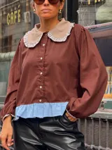 Camisa de color marrón con cuello Peter Pan en tono beige con borde de encaje, mangas largas abullonadas con puños elásticos y detalle de volado en el ruedo en color celeste.