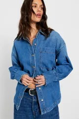 Camisa de denim azul de calce amplio, con cuello clásico, botonadura frontal, bolsillo plaqué en el pecho, hombros caídos y puños anchos abotonados.