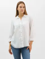 Camisa de tencel color blanco, con cuello camisero, cierre frontal de botones y dos bolsillos en el pecho. Presenta mangas largas con presilla para regular el largo y ruedo con detalle de flecos.