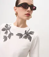 Remera blanca de manga corta y cuello redondo, confeccionada en algodón con bordados florales en tono oscuro aplicados en los hombros.