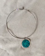 Gargantilla rígida de metal plateado con dije circular central que presenta una piedra turquesa con vetas oscuras enmarcada en una base metálica.