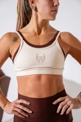 Top deportivo de doble capa en color crema con detalles en marrón oscuro, con breteles finos y logo bordado en el centro del pecho.