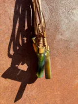 Collar con dije cilíndrico de resina verde con inclusiones blancas, engarzado en metal dorado y colgante de cordón de cuero marrón.