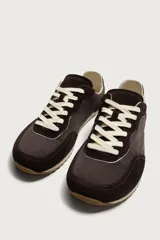Zapatilla deportiva estilo barefoot, color marrón, con diseño de paneles en combinación de materiales y detalles en serraje. Cuenta con puntera redondeada y ancha, cordones en color crema y suela fina de goma flexible en contraste.