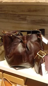 Bolso tipo tote de cuero marrón con acabado brillante, diseño estructurado con detalles de remaches metálicos en las asas y cierre de cordón ajustable.