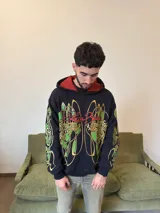 Hoodie oversize de color negro con estampado gráfico en pecho y mangas en tonos amarillos y verdes. Presenta un forro interior de capucha en color rojo con textura tejida y logo bordado en la manga.