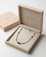 Collar de acero con baño de oro, compuesto por una combinación de perlas naturales blancas, piedras semipreciosas en tonos verdes y turquesas, y pequeñas cuentas doradas. Presenta un dije central de piedra verde oscura y un cierre con cadena de alargue que incluye una medalla grabada con las iniciales MA.