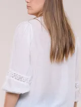 Blusa blanca de manga larga con escote en V y detalles de encaje en la botonera frontal, cuello y puños.