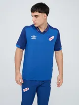 Remera polo deportiva Umbro del Club Nacional de Football, color azul con cuello en V y detalles en rojo y blanco. Incluye el logo de Umbro y el escudo del club.
