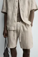 Bermuda de lino color blanco hueso, de corte relajado y tiro medio. Presenta cintura elástica ajustable con cordón, pliegues frontales y bolsillos laterales y trasero.